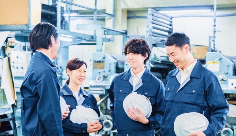 工場内で作業服を着てヘルメットを持ち、笑顔で会話する4人の従業員